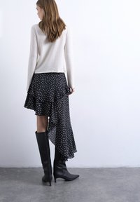 Γυναίκα με μακριά μαλλιά που φοράει λευκό πουλόβερ, μαύρη ασύμμετρη φούστα με πουά και μαύρες μπότες με τακούνι μέχρι το γόνατο, στέκεται σε γκρι πάτωμα.