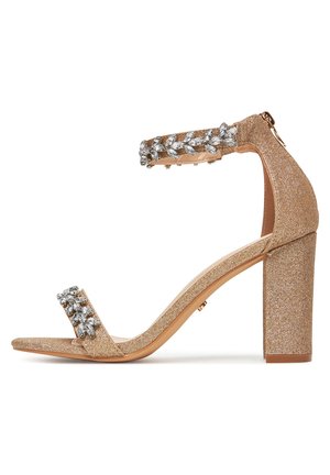 Sandalo con tacco alto a blocco color oro glitterato, con cinturino alla caviglia e cristalli decorativi sul cinturino. Design a punta aperta. Texture liscia.