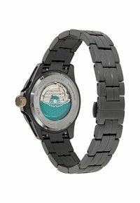 Černé metalové hodinky na zápěstí s náramkem na články, vitrínové dno s tyrkysovými detaily a stříbrným mechanickým strojkem.