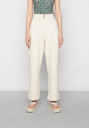 Kremowe spodnie z wysokim stanem i plisami, z dwoma guzikami w pasie, noszone z białymi sandałami na platformie i zielonym, kwiecistym, bezrękawnym topem.
