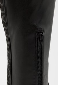 Bota de cuero negro hasta la rodilla con cierre lateral, patrón texturizado y logo de Buffalo en el lado exterior. Acabado mate, diseño robusto.