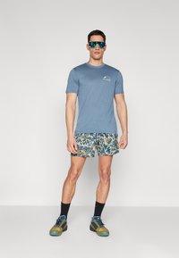 Men's casual outfit featuring a light blue short-sleeve shirt, patterned swim shorts, and colorful athletic shoes with dark socks.