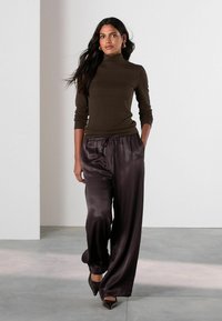Brown turtleneck sweater with long sleeves paired with dark satin wide-leg pants. Model wearing pointed-toe heels, standing in a minimalist setting.