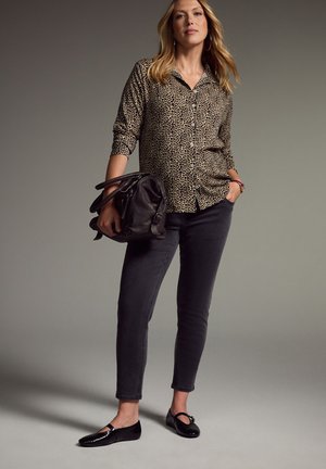 Kvinde iført leopardmønstret skjorte med knapper, sorte jeans og sorte ballerinasko, der holder en stor sort håndtaske med den ene hånd i lommen.