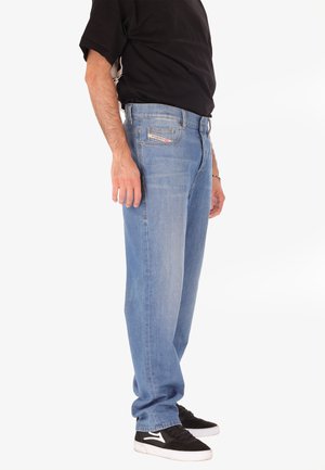 Homme portant un jean bleu clair, un t-shirt noir et des baskets noires avec des semelles blanches, se tenant de côté sur un fond blanc.