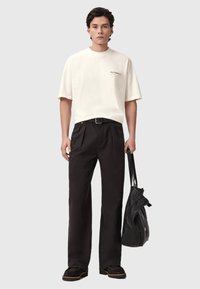 Μπεζ κοντομάνικη μπλούζα με το λογότυπο "ALL-SAINTS", σε συνδυασμό με μαύρο ψηλόμεσο παντελόνι και μαύρα παπούτσια. Το μοντέλο κρατά μαύρη τσάντα.