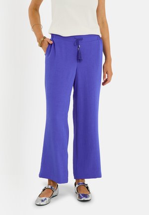 Femme portant un pantalon large bleu vif avec un cordon de serrage, un haut blanc sans manches, des chaussures Mary Jane argentées et des bracelets dorés à un bras.