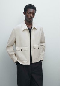 Beige, letvægtsjakke med krave, to forlommer med sorte knapper og lynlås. Bæres over en sort V-hals trøje.