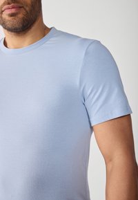 Tricou scurt, de culoare albastru deschis, dintr-un material neted și elastic, cu un guler clasic rotund și detalii discrete ale cusăturii.