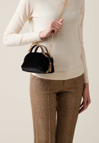 Black velvet handbag with gold accents, zip closure, and braided handles, worn crossbody over a light sweater and patterned pants.