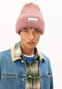 Roze gebreide beanie met een label, gedragen met een spijkerjack en een geruit overhemd. De beanie heeft een zachte textuur en een gestructureerde vorm.