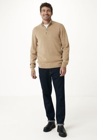 Béžový svetr s krátkým zipem vyrobený z měkké pleteniny, kombinovaný s tmavými džínami a černými kotníkovými botami, s ohrnutými manžetami a volným střihem.