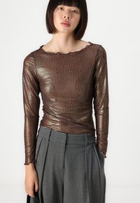 Shiny, brown long-sleeve top with a leopard print pattern and a scalloped neckline, paired with pleated gray trousers.