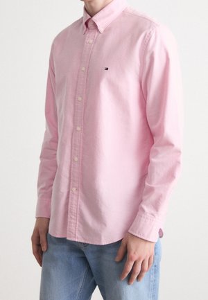Man wearing long-sleeve pink button-up shirt with small logo on chest, paired with light blue jeans against plain white background.
