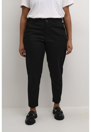Person trägt schwarze, schmal zulaufende Hose mit Knopf und Reißverschluss, kombiniert mit schwarzen Loafers und einem weißen Kurzarmhemd.