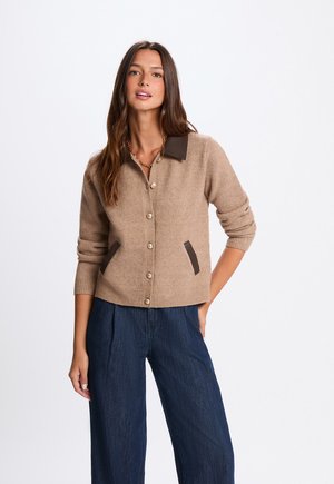 Woman wearing a beige buttoned cardigan with dark collar and pocket details, paired with dark blue wide-leg pants.