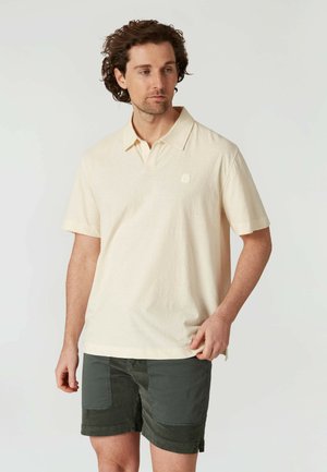 Lys beige kortærmet polo-shirt med krave og struktureret stof; har et lille logo på brystet og passer godt sammen med mørkegrønne shorts.
