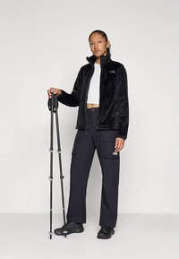 Črna flis jakna, bela srajca in črne hlače s številnimi žepi. Drži pohodne palice. Mat tekstura, funkcionalna zasnova, pripravljeno na aktivnosti na prostem.