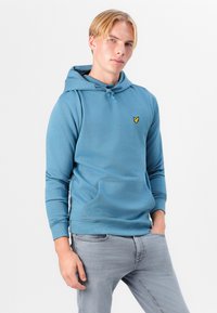 Svetlomodrá mikina s kapucňou a klokaním vreckom, s nastaviteľnou šnúrkou na kapucni, rebrovanými manžetami a malým žltým logom na hrudi.