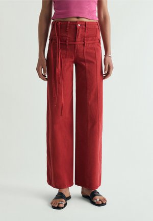 Mujer con pantalones rojos de talle alto y pierna ancha con detalles de costura frontal y cinturón de tela a juego, combinados con un top corto rosa y sandalias negras.