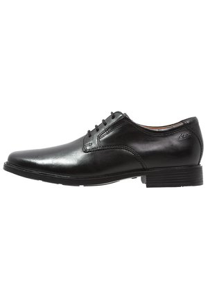 Chaussure habillée en cuir noir avec un bout arrondi, texture lisse, six œillets pour les lacets et une semelle en caoutchouc basse.