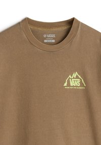 Camiseta marrón de corte suelto con cuello acanalado. Presenta un logo de montaña verde con el texto "VANS" y "HECHA PARA LOS ELEMENTOS". Material de algodón.