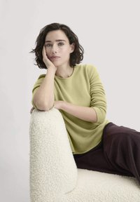 Grüner Strickpullover mit langen Ärmeln und rundem Ausschnitt, kombiniert mit dunklen Hosen, sitzt auf einem texturierten weißen Stuhl. Neutraler Hintergrund.