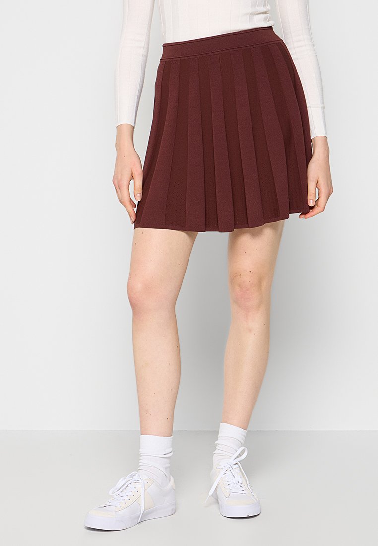 Burgunderfarbene plissierte Rock mit hohem Bund, aus strukturiertem Stoff, kombiniert mit weißen Sneakers und Socken, getragen von einem Modell.