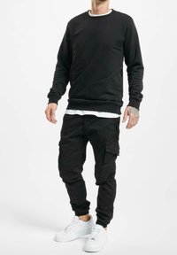 Sweat-shirt en polaire noir à col rond, associé à un pantalon cargo noir avec des poches latérales. Une paire de baskets blanches complète la tenue.