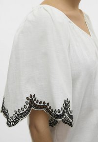 Blusa bianca con maniche corte e a sbuffo. Presenta un delicato bordo ricamato nero lungo i bordi delle maniche e un tessuto leggero e con texture.