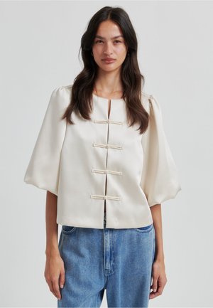 Kermanvärinen lyhyt pusero, jossa on puffihihat, avainreikäkaulus ja neljä kangasnapillista suljinaa. Yhdistetty sinisiin korkeavyötäröisiin farkkuihin.