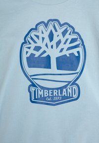 Lyseblå genser med en stor blå Timberland-logo med et tre-design og teksten "Est. 1973". Myk tekstur, rund hals.