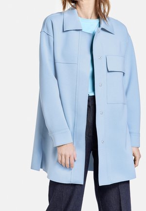 Personne portant une surchemise oversize bleu clair avec une poche poitrine à rabat, associée à un pantalon foncé et un top bleu clair en dessous.