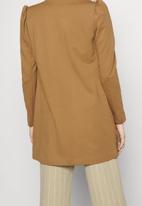 Hnědá tunika s dlouhým rukávem a designem s vycpanými rameny; jemná tkanina; v kombinaci s béžovými kalhotami s proužky. Zobrazeno zezadu.