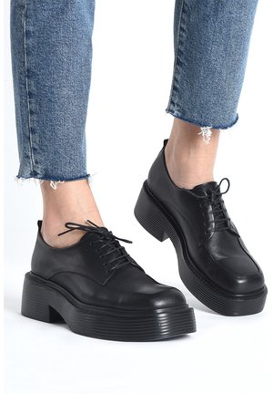 Zapatos de cuero negro con punta redonda, diseño con cordones y suela gruesa con surcos. Llevados con unos vaqueros de denim azul cortos con dobladillos deshilachados.