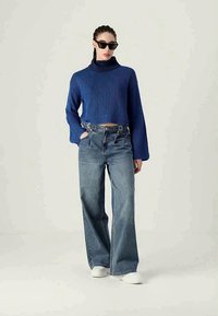 Suéter cropped de malha azul com gola alta, combinado com jeans de algodão de cintura alta e corte largo, e sapatilhas brancas. Óculos de sol pretos usados.