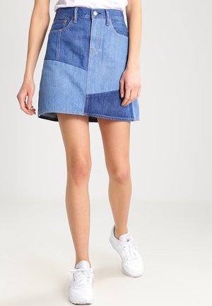 Femme portant une mini-jupe en jean patchwork avec différentes nuances de bleu, des baskets blanches et un haut blanc, debout devant un fond uni.