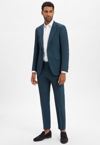 Türkisfarbener maßgeschneiderter Anzug aus glattem Stoff, mit einem Ein-Knopf-Verschluss und schlanken Hosen, kombiniert mit einem weißen Dress-Shirt und schwarzen Slippern.