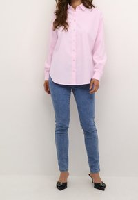 Chemise rose pâle à manches longues avec boutons et col pointu, associée à un jean skinny bleu et des talons noirs à bout pointu.