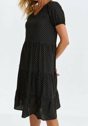 Kvinde iført en sort knælang kjole med korte puffede ærmer og små hvide polkaprikker, stående mod en ensfarvet baggrund.