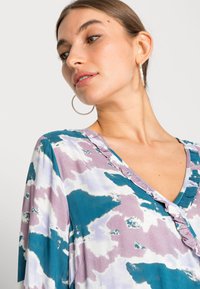 Blusa fluida com decote em V, adornada com padrões pastel em teal, roxo e branco; destacada por um acabamento em babados ao longo do decote.