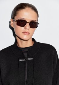 Occhiali da sole rettangolari neri con lenti scure, indossati con una giacca in maglia nera caratterizzata da un colletto alto e un dettaglio di chiusura frontale.