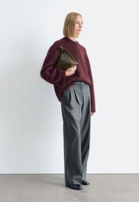 Weinrote, kuschelige Pullover kombiniert mit grauen, plissierten Hosen. Das Model hält eine dunkle, geflochtene Handtasche und trägt marineblaue Schuhe vor einem neutralen Hintergrund.