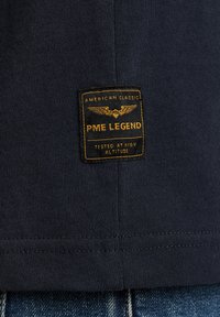 Navyblauwe katoenen t-shirt met een gestikt label waarop "PME Legend" staat, met gele tekst en een ontwerp van een gevleugeld embleem, met ribbelstructuur.