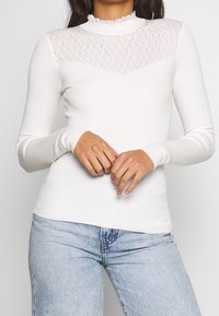 Vit ribbad långärmad topp med en genomskinlig spetsdetalj vid axlarna, med en mock neck och volangkant. Kombination med ljusblå denimjeans.