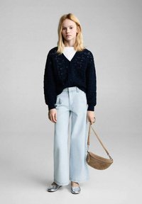 Cardigan in maglia blu navy con collo a V, jeans a gamba larga blu chiaro e scarpe basse argentate; tiene una piccola borsa a tracolla in suede color tan.