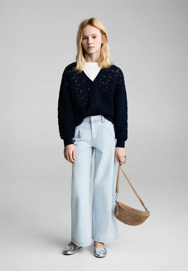 Cardigan in maglia blu navy con collo a V, jeans a gamba larga blu chiaro e scarpe basse argentate; tiene una piccola borsa a tracolla in suede color tan.