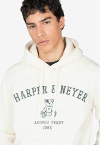 Sudadera color crema con cordones, que presenta un gráfico desgastado de un oso de peluche y el texto "HARPER & NEYER ARTHUR TEDDY 1985" en verde oscuro.