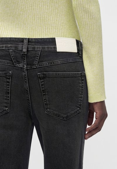 Άτομο που φοράει μαύρο τζιν από denim με ένα λευκό ορθογώνιο ετικέτα που γράφει "CLOSED" και ένα ανοιχτό πράσινο ριμπ πλεκτό πουλόβερ, κομμένο στη μέση.
