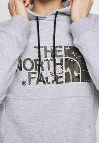 Personne portant un sweat à capuche gris clair avec des cordons noirs et un logo camouflage "THE NORTH FACE" sur la poitrine, les mains ajustant la capuche.